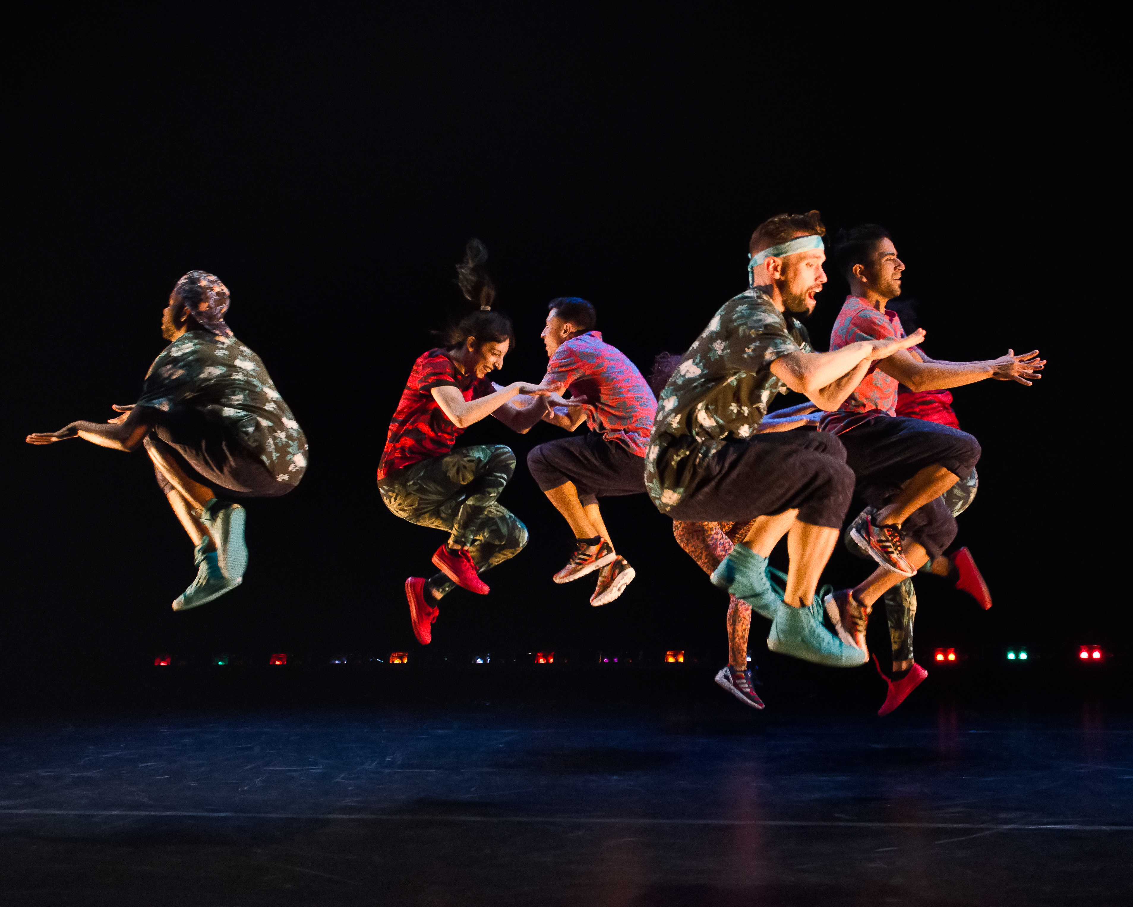 7 dancers in colorful clothing jump in unison.