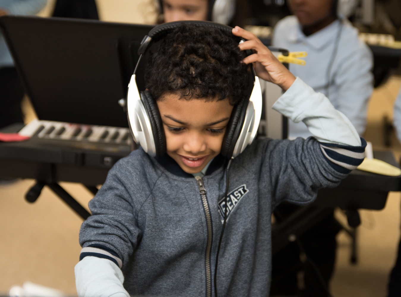 An adorable young boy grasps his oversized headphones with one hand as he smiles with joy while he plays piano.