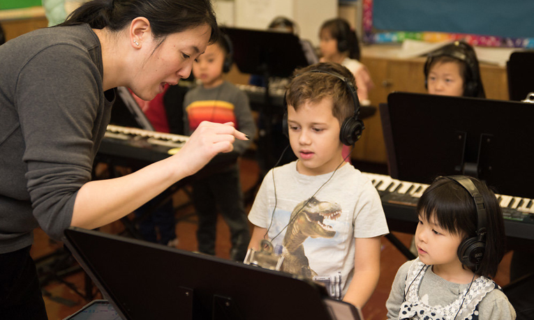 Two children are wearing headphones and sharing a piano. They are reading the music while their teacher helps them keep a steady beat.