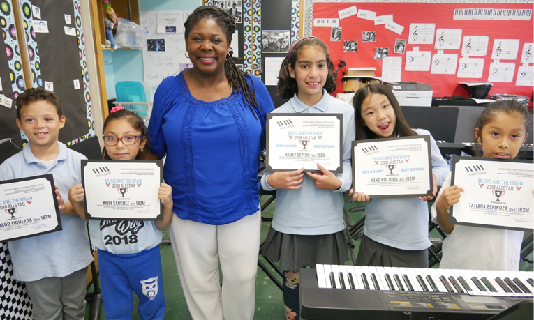 A smiling teacher stands with her arms around five of her students. Each child is displaying an All Star achievement certificate.