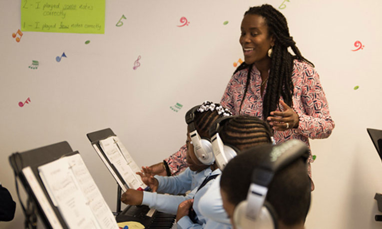 A teacher is smiling as she sings along with her students as they play the piano in class.