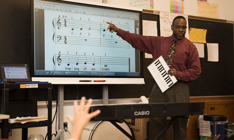 A teacher gestures to sheet music on an interactive whiteboard during a lesson.