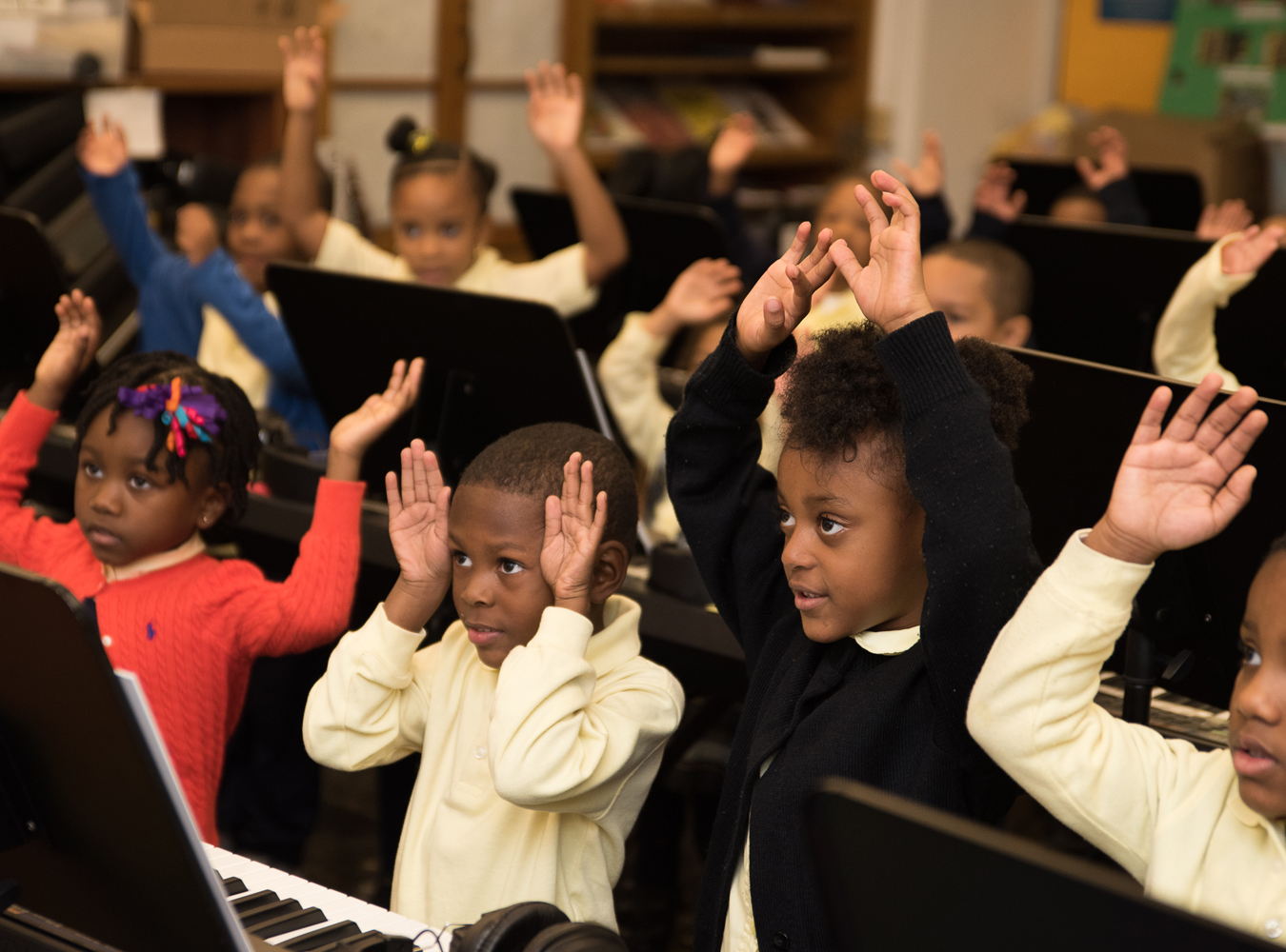 Young children stand at their pianos stretching their arms up in the air as they follow their teacher's lead.