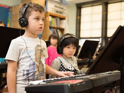 A young boy and a young girl stand together at a piano in class.