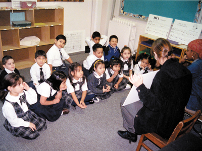 A photo from the early 90s shows two teachers working with a class of young children. The teachers are showing the children a poster of a song.,