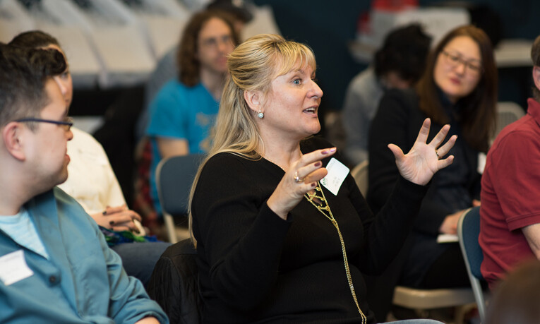 A music teacher gestures with her hands while addressing a room of her peers.