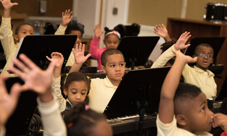 A class of students stands at their pianos. They all have their hands in the air ready to answer their teacher's question.