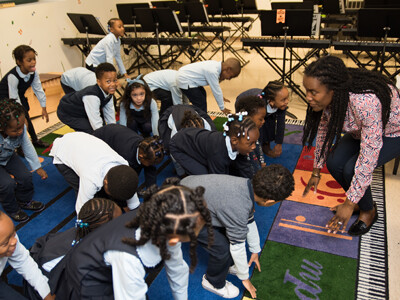 Young children copy their teacher while they are learning the dance to a new song. Everybody has their hands on the ground. They are looking at each other, smiling and laughing.