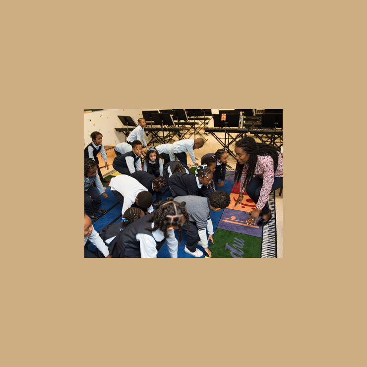 Young children copy their teacher while they are learning the dance to a new song. Everybody has their hands on the ground. They are looking at each other, smiling and laughing.